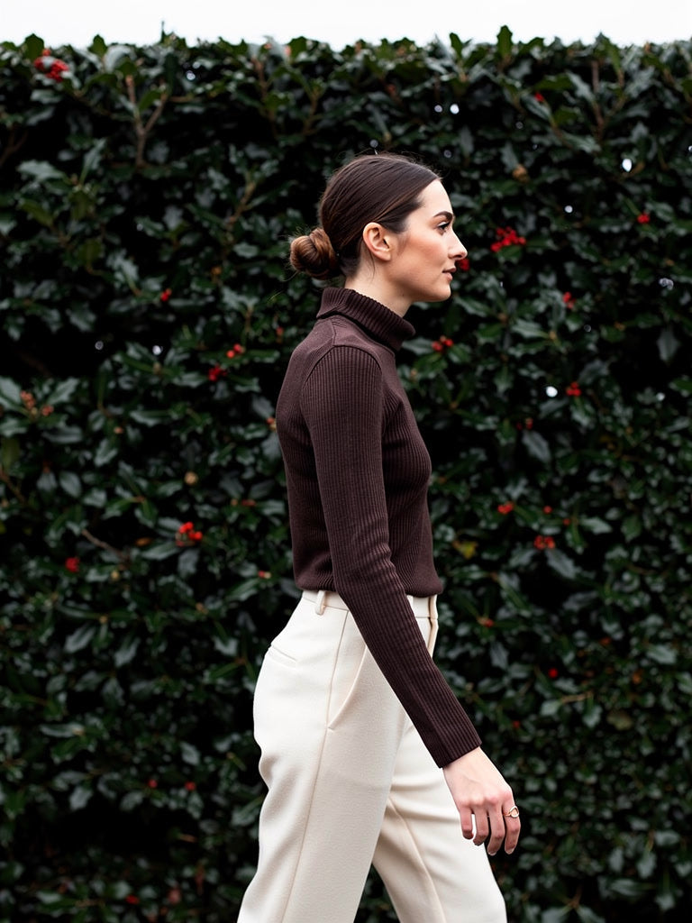 Wool roll neck top in treacle brown worn in profile against a winter holly hedge
