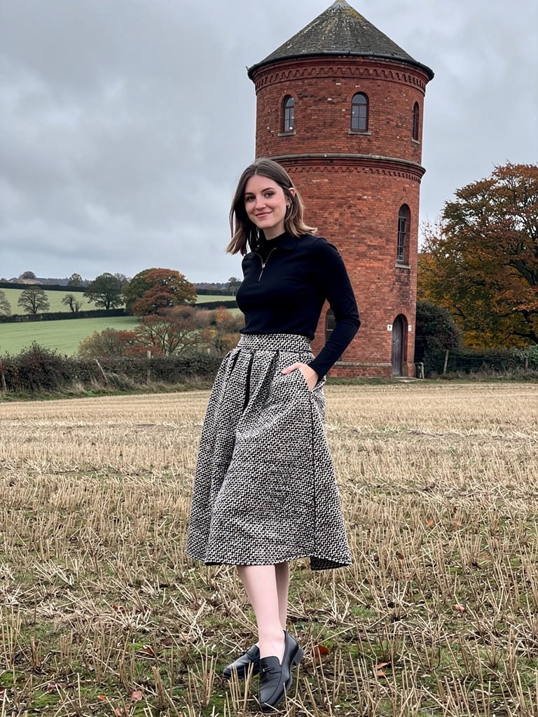 Black and white herringbone A line skirt worn with a black polo neck and flat loafers, photographed beside a Victorian brick water tower