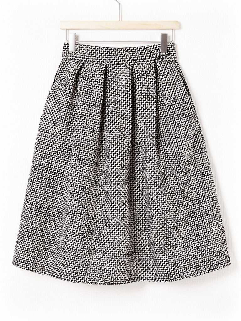 Black and white herringbone A line skirt on a wooden hanger, showing the wide plain waistband, the deep soft pleats and the level knee length hem