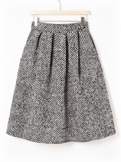 Black and white herringbone A line skirt on a wooden hanger, showing the wide plain waistband, the deep soft pleats and the level knee length hem