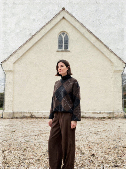 Womens argyle jumper in chocolate brown and black worn with wide brown trousers outside a village chapel