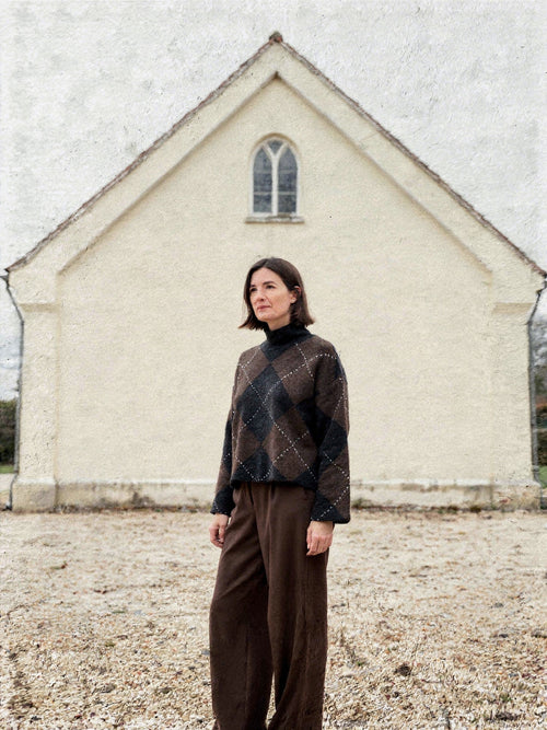 Womens argyle jumper in chocolate brown and black worn with wide brown trousers outside a village chapel
