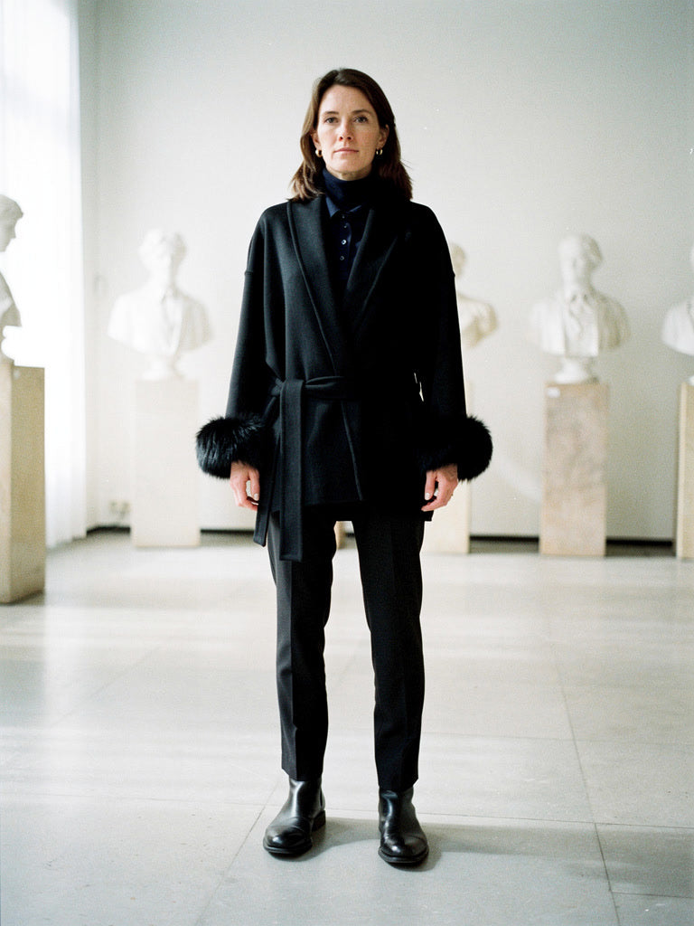 Woman in a black belted coat with faux fur cuffs in a sculpture gallery