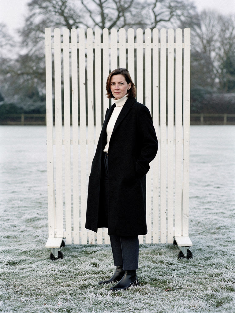 Woman wearing a black coat open over a cream roll neck on a frosted cricket ground