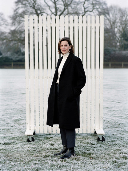 Woman wearing a black coat open over a cream roll neck on a frosted cricket ground