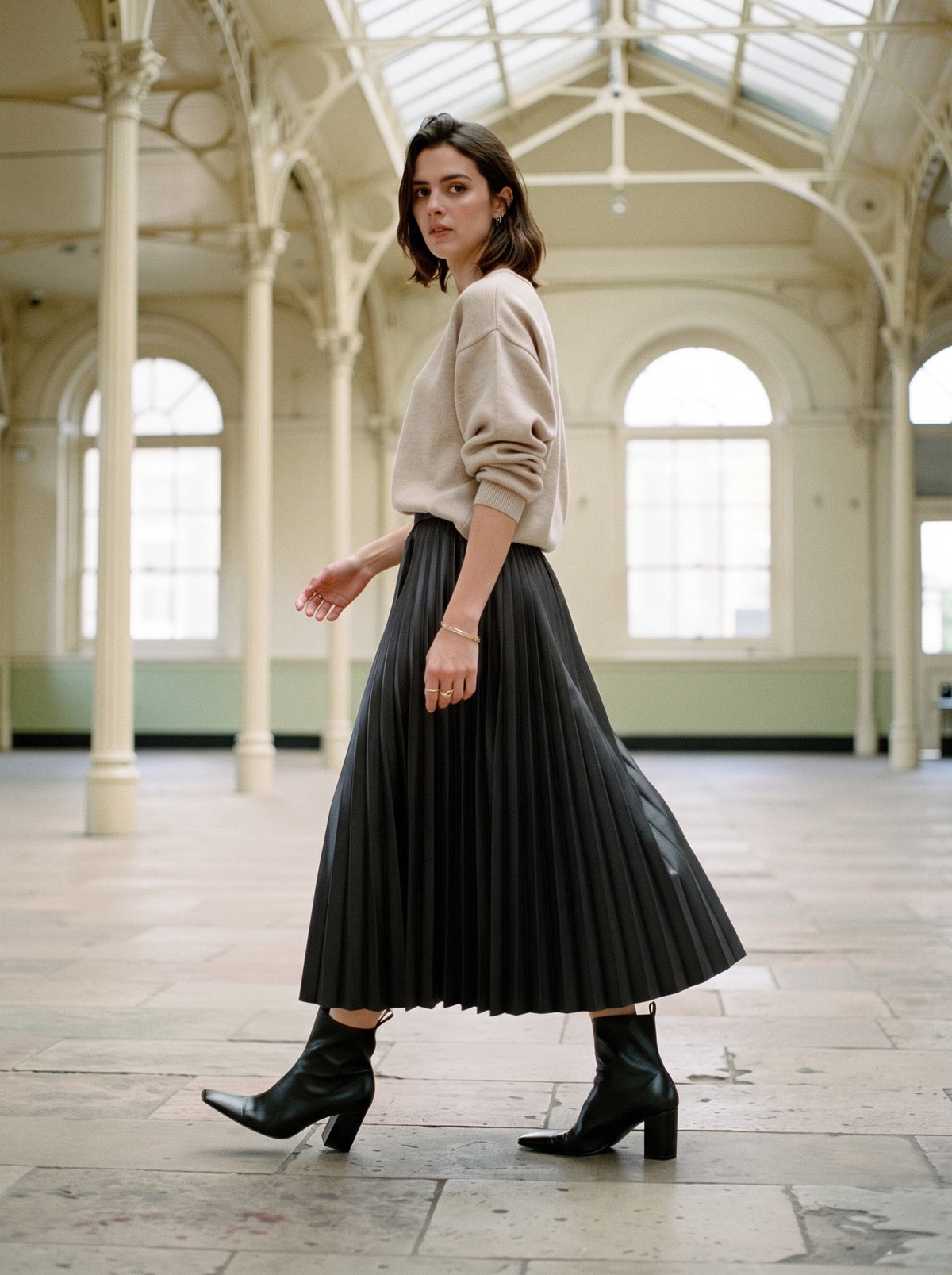 Woman walking in a black pleated skirt in faux leather, Rook, worn with a fine oatmeal knit and black ankle boots in an empty Victorian corn exchange