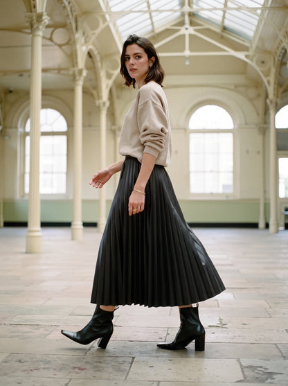 Woman walking in a black pleated skirt in faux leather, Rook, worn with a fine oatmeal knit and black ankle boots in an empty Victorian corn exchange