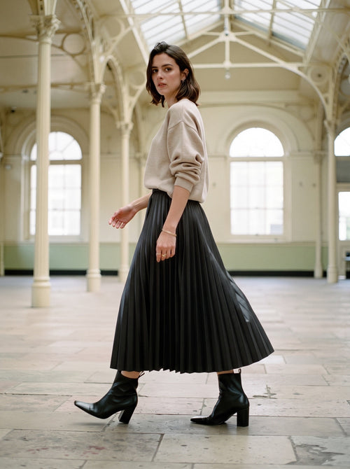 Woman walking in a black pleated skirt in faux leather, Rook, worn with a fine oatmeal knit and black ankle boots in an empty Victorian corn exchange