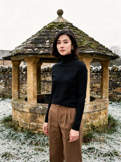 Woman wearing a black roll neck jumper beside a stone village well house in winter