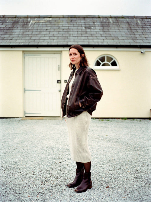 Womens brown jacket in distressed faux leather, worn as an oversized bomber with a knitted midi skirt in a coach house yard