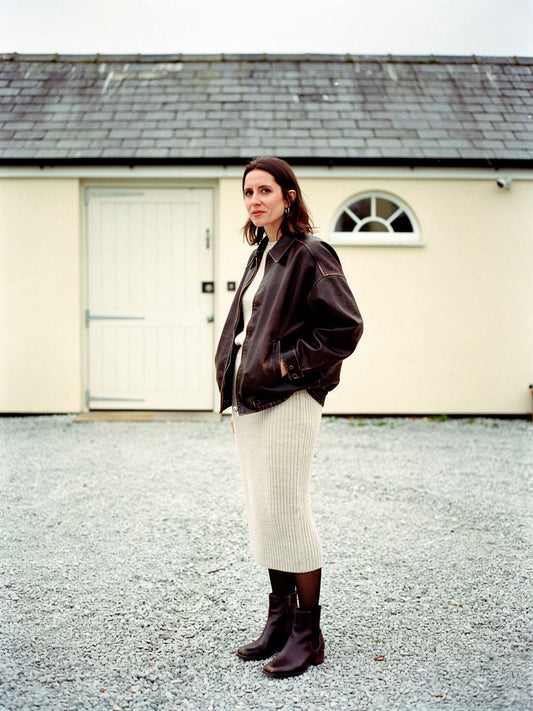 Womens brown jacket in distressed faux leather, worn as an oversized bomber with a knitted midi skirt in a coach house yard