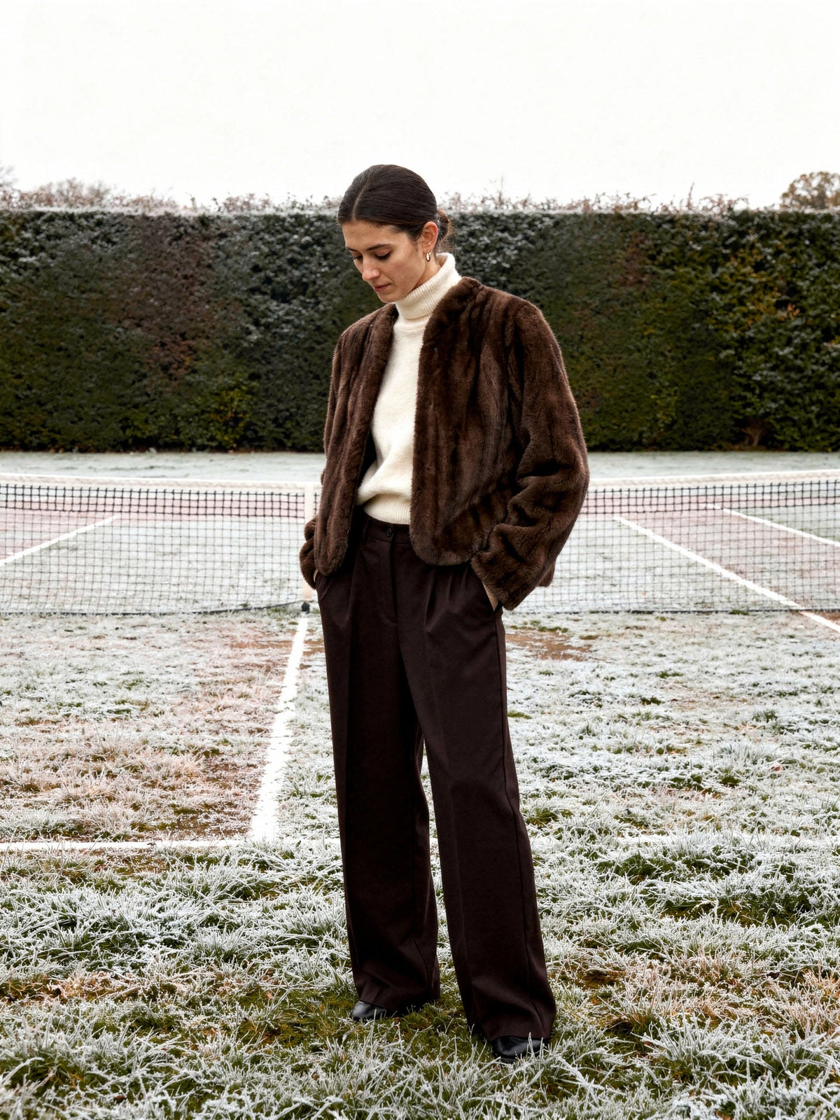 Woman in a brown faux fur jacket standing at the edge of a frosted grass tennis court in midwinter