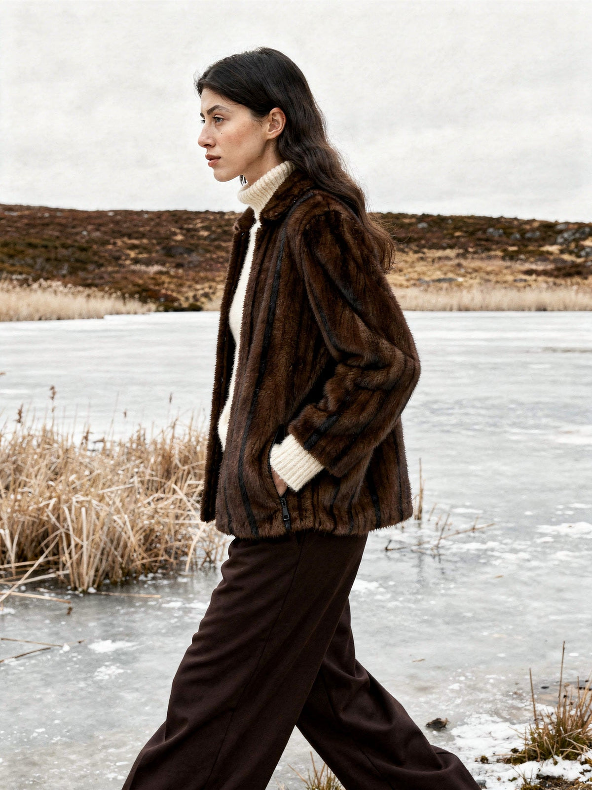 Brown faux fur jacket worn open and seen in profile beside a frozen upland tarn