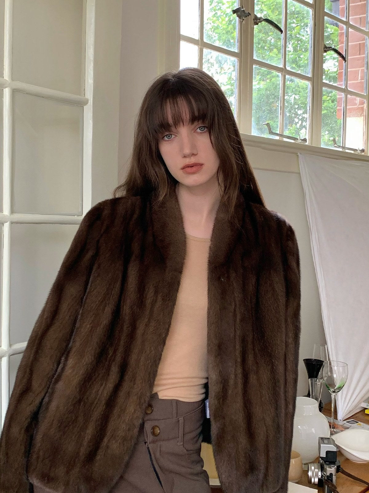 Brown faux fur jacket worn open over a fine knit, photographed indoors beside a window