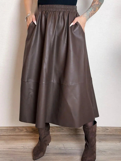 Maker photograph of the brown faux leather skirt worn, both hands in the slanted side pockets, showing the gathered waistband and the level seam low on the skirt