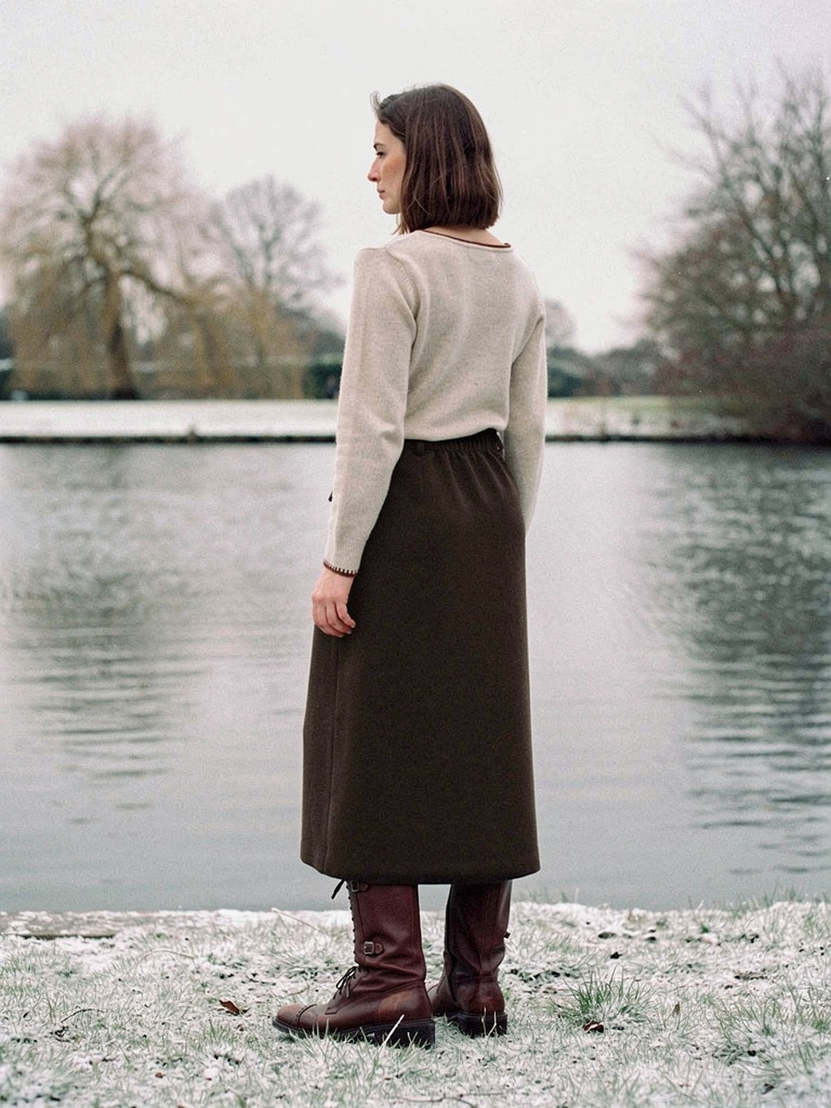 Brown skirt in a brushed A line midi cut, seen from behind beside a village pond, in Bog Oak