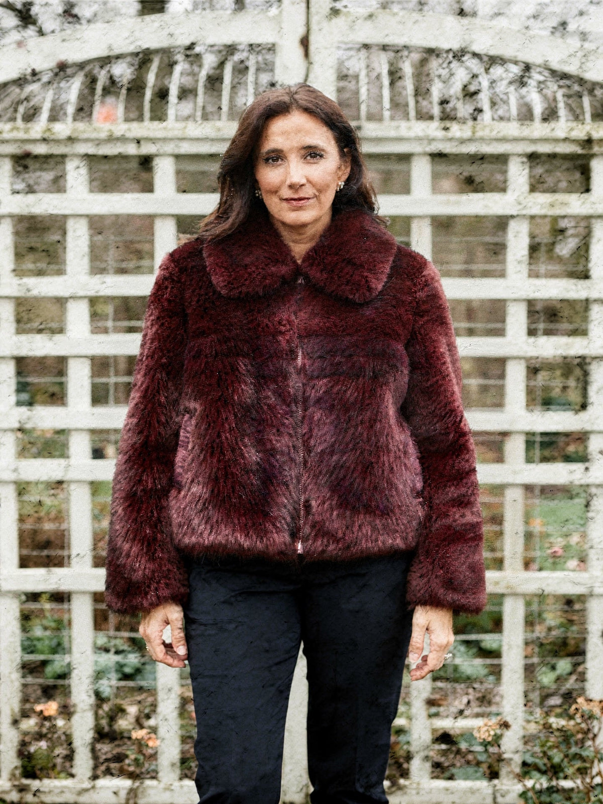 Woman wearing a burgundy faux fur jacket zipped to the neck in front of a white garden aviary
