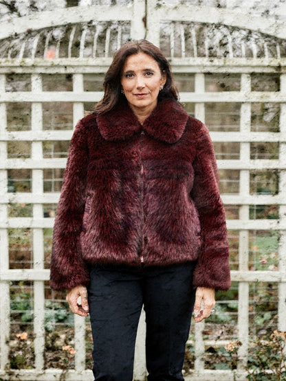 Woman wearing a burgundy faux fur jacket zipped to the neck in front of a white garden aviary