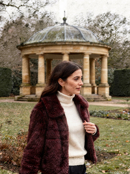 Burgundy faux fur jacket worn open over a cream roll neck beside a stone belvedere