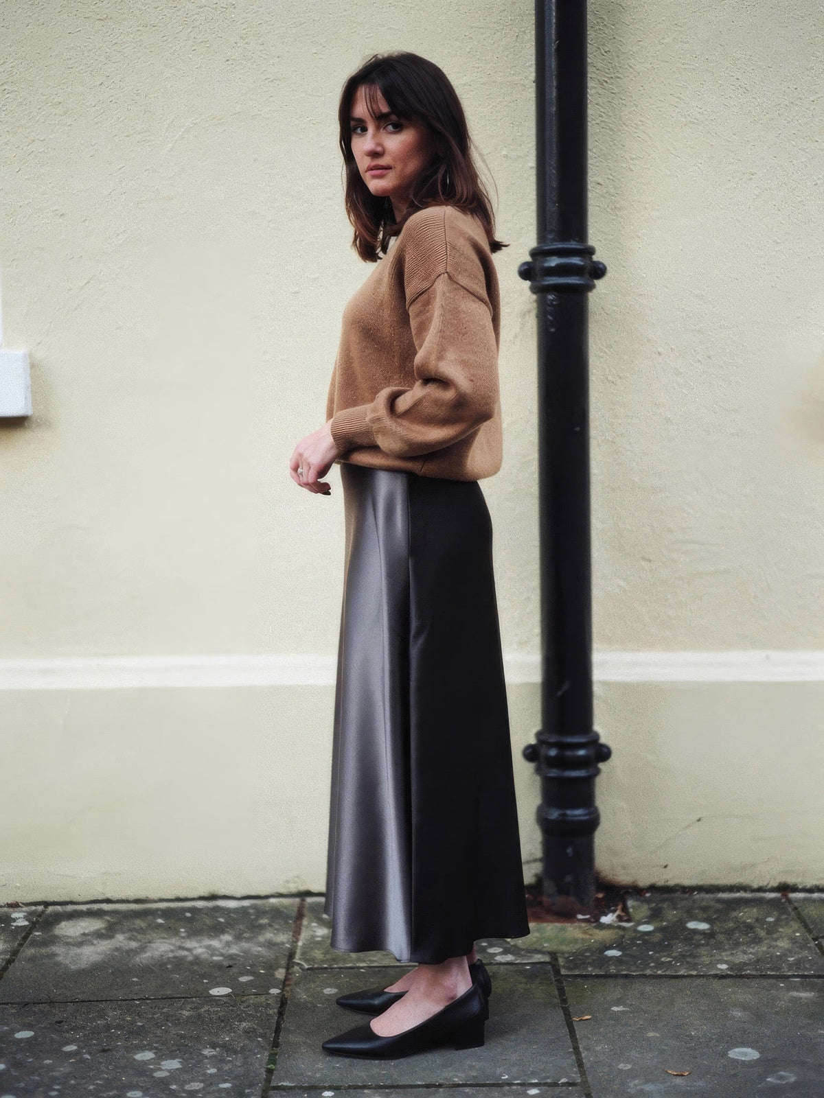 Black satin skirt worn with a camel jumper against a pale stucco wall, showing the ankle length hem and the quiet lustre of the satin