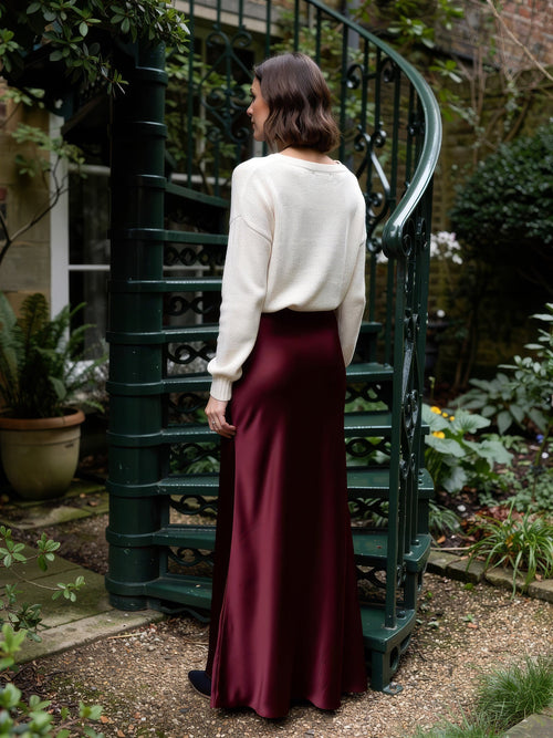 Burgundy satin skirt in Malbec wine red worn with a cream jumper, seen from behind at the foot of a cast iron spiral staircase in a walled town garden, showing the full ankle length sweep
