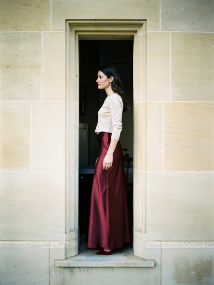 Burgundy satin skirt in Malbec wine red worn in profile in a pale limestone doorway, showing how the bias cut satin falls from waist to ankle