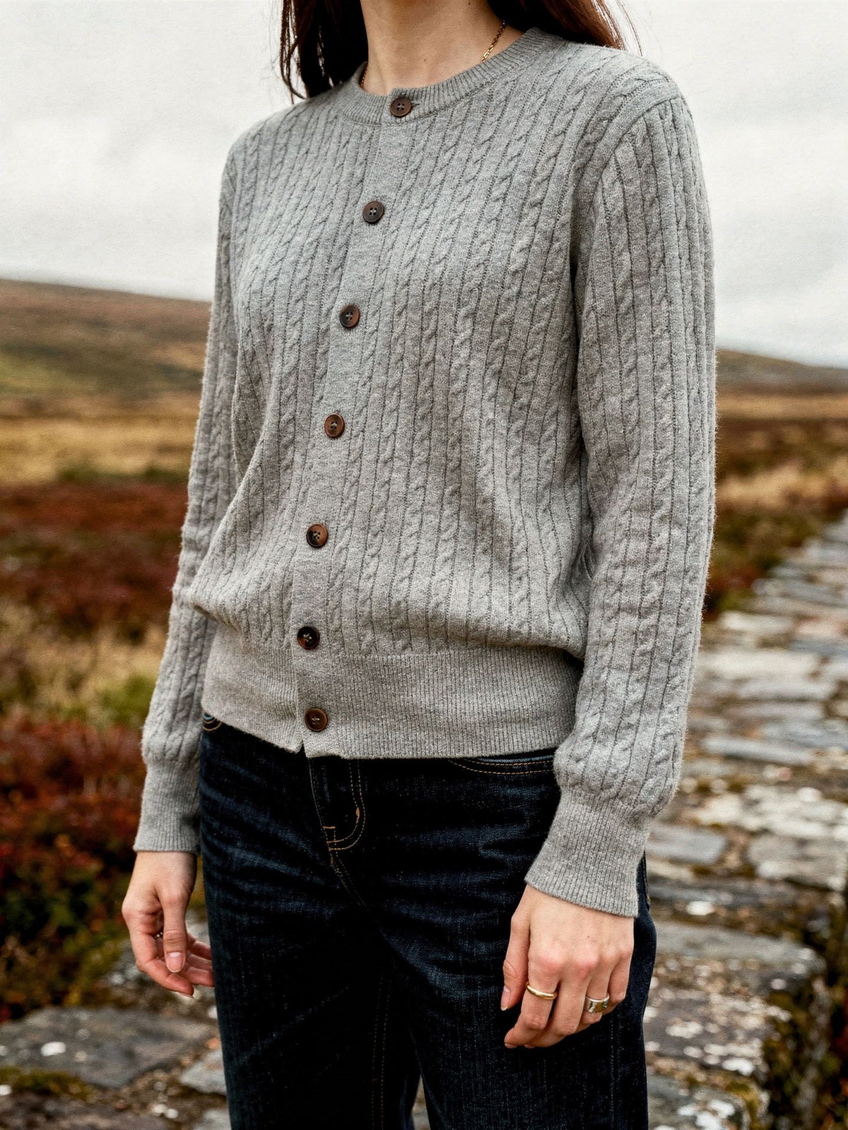 Womens cable knit cardigan in Harbour Grey, worn with dark jeans on a moorland bridge