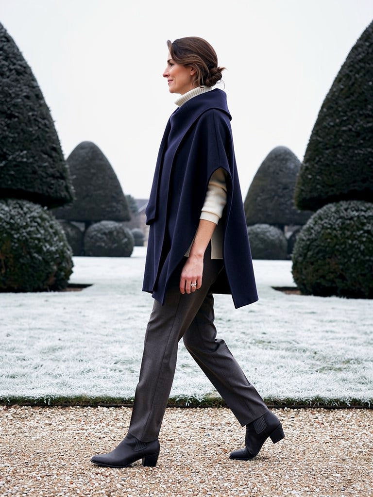 Woman walking in profile through a frosted topiary garden wearing the Ensign navy cape coat