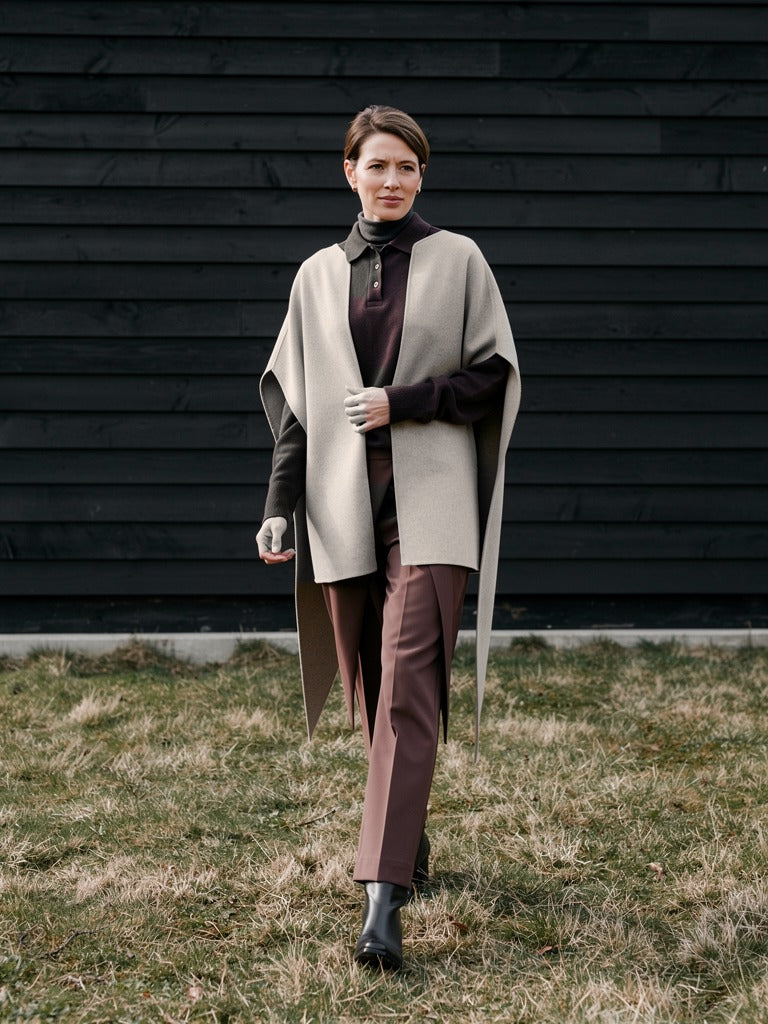 Woman walking past a black tarred barn wall wearing the Tussock cape coat open over a brown polo neck
