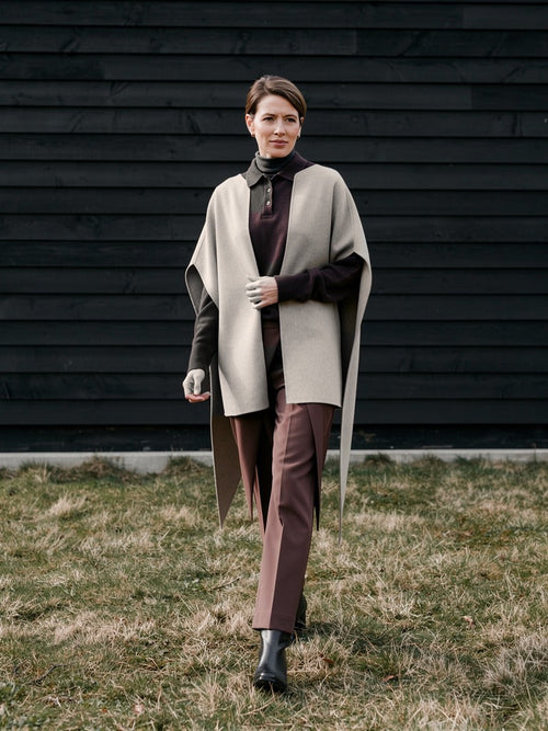 Woman walking past a black tarred barn wall wearing the Tussock cape coat open over a brown polo neck