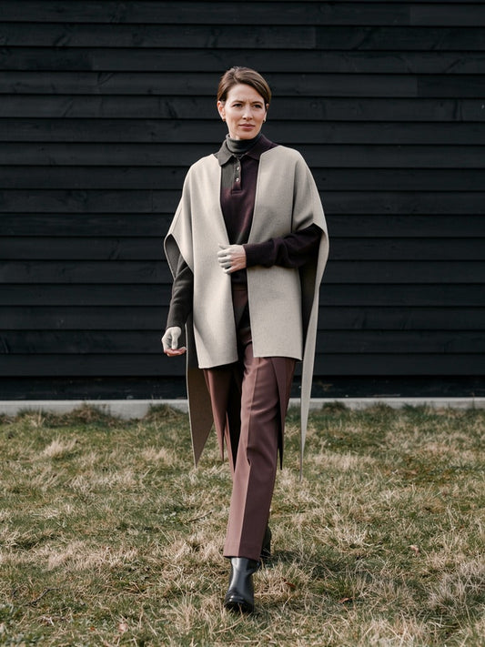 Woman walking past a black tarred barn wall wearing the Tussock cape coat open over a brown polo neck