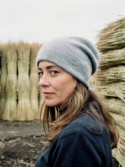 Woman wearing a grey cashmere beanie in a thatcher's yard, stacked reed bundles behind her