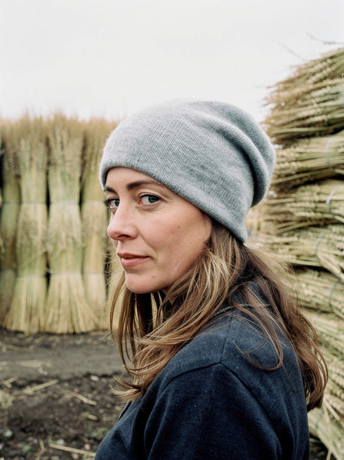 Woman wearing a grey cashmere beanie in a thatcher's yard, stacked reed bundles behind her