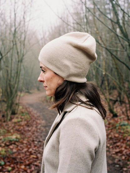 Woman in profile wearing a malt cashmere beanie turned up at the brow, in a hazel coppice