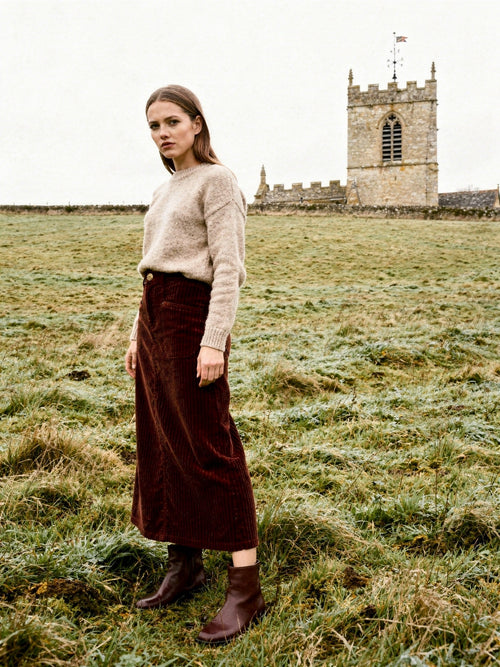 Woman wearing a long corduroy skirt in Amarone burgundy in a frosted winter meadow, the square stone tower of a village church rising behind her