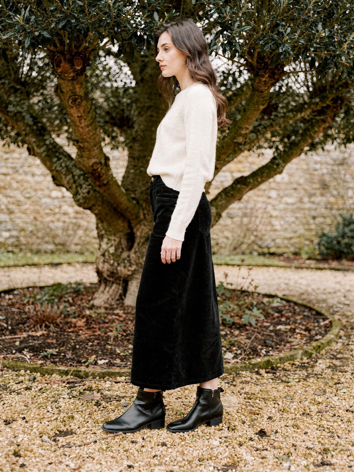 Woman wearing a long corduroy skirt in Sloe black in a walled garden, seen in profile, the hem falling on the lower calf above black ankle boots