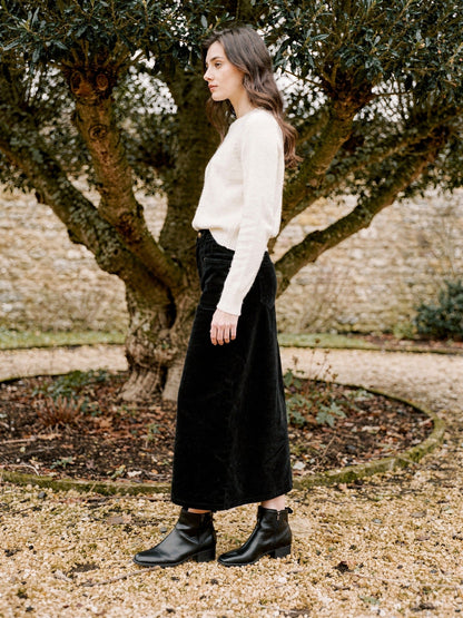 Woman wearing a long corduroy skirt in Sloe black in a walled garden, seen in profile, the hem falling on the lower calf above black ankle boots