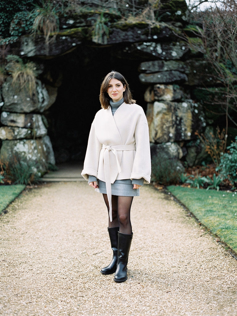 Womens cream collarless wrap jacket in Eggshell, belted at the waist, worn in front of a stone grotto