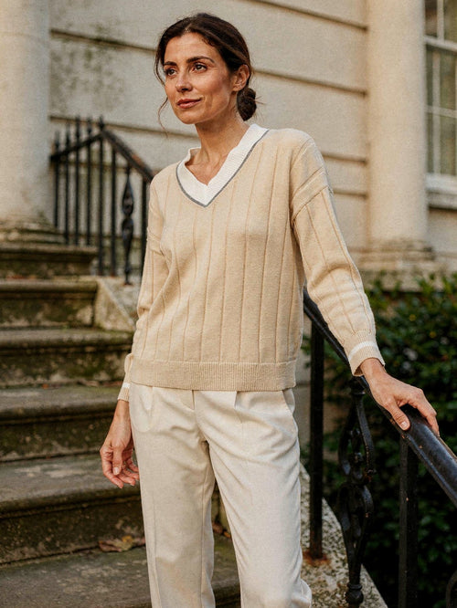 Woman wearing the stone V-neck cricket jumper in cotton, with contrast ivory collar, on townhouse steps