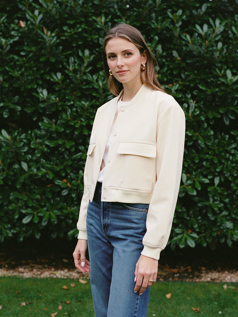 Woman wearing a cropped bomber jacket in buttermilk cream, open over a white t-shirt and high waisted jeans, in front of a dark laurel hedge