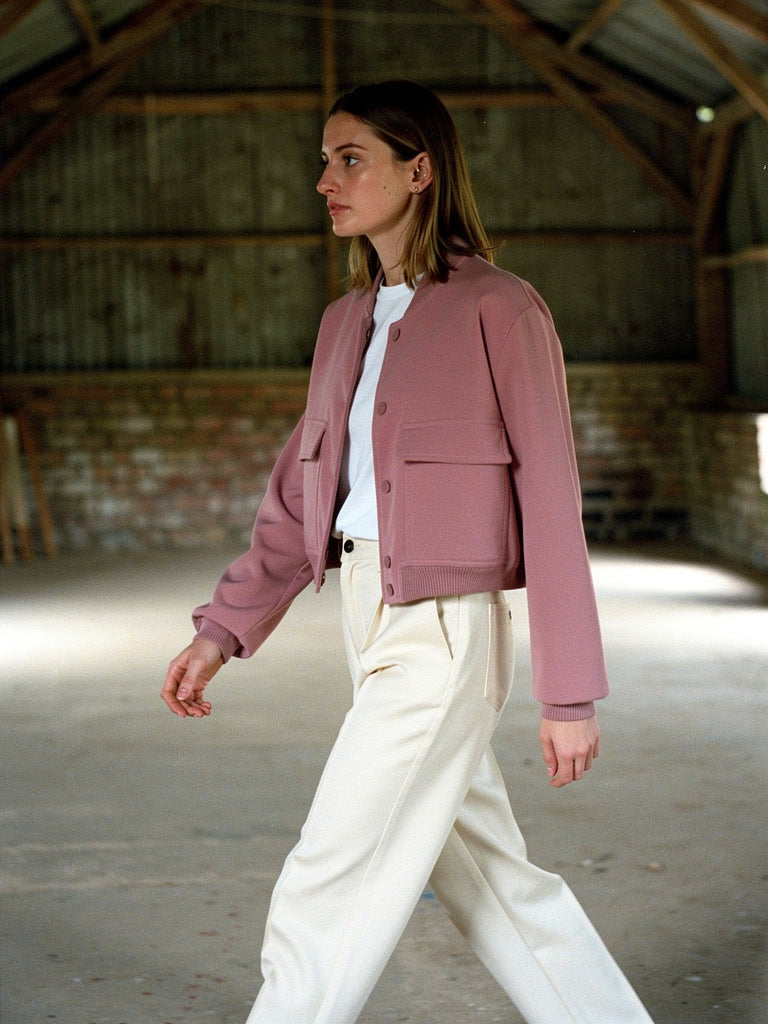 Woman walking in a cropped bomber jacket in dusty rose, worn open over cream trousers in an empty barn loft