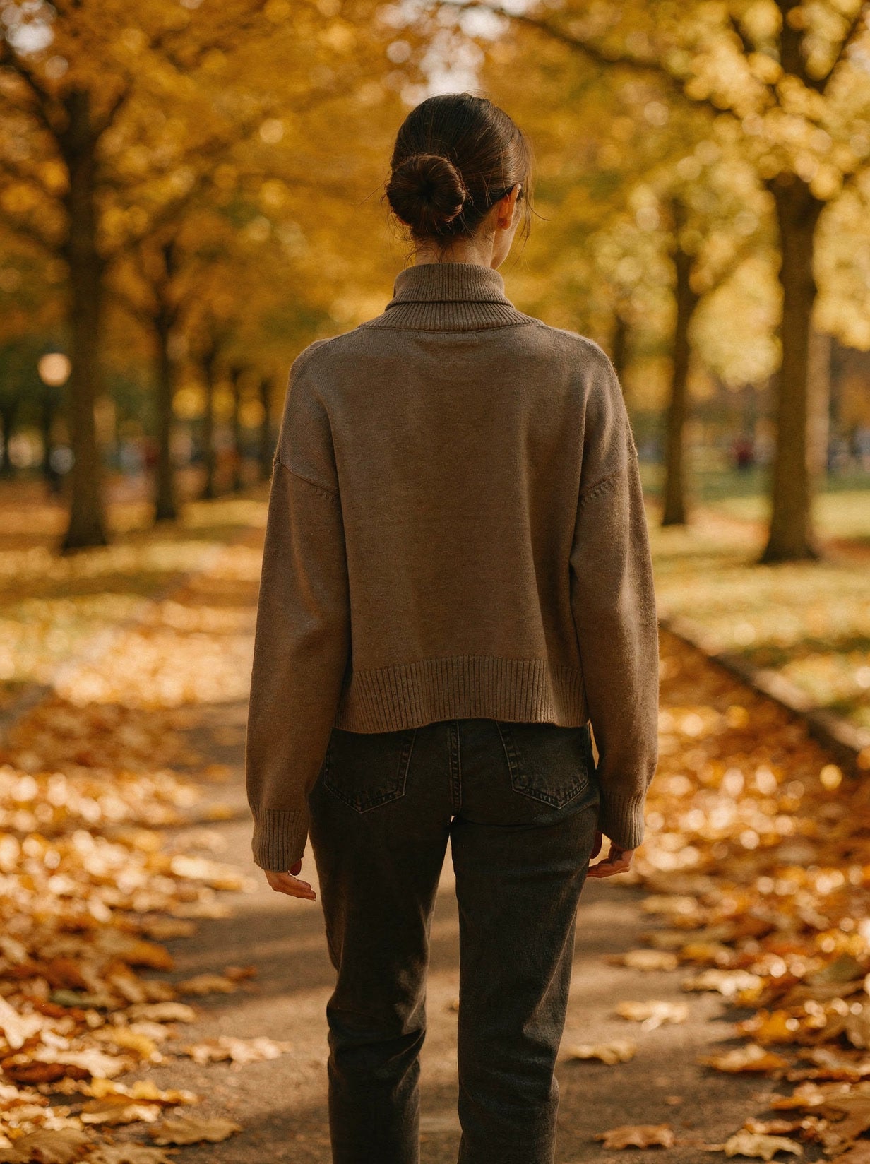 Back view of the taupe cropped turtleneck jumper showing the dipped high-low hem, worn in an autumn park