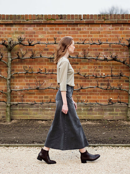 Charcoal grey denim maxi skirt worn in profile beside an espaliered brick garden wall