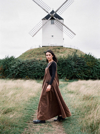 Woman wearing a Rust Wash denim pinafore dress, washed A-line maxi, by an English windmill
