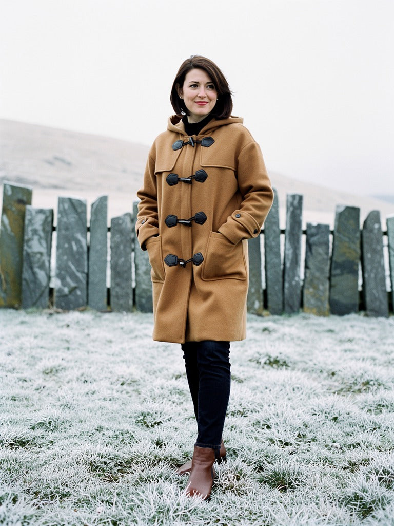 Woman wearing a camel duffle coat with the hood down and the toggles fastened, beside a slate slab fence