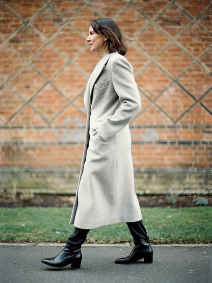 Fitted longline coat in Pumice grey worn walking past a red brick wall, ankle length with a nipped waist