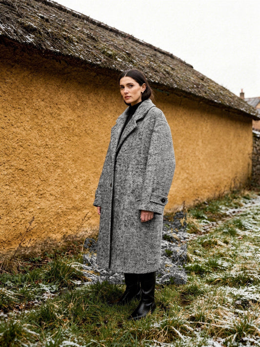 Woman wearing a grey herringbone midi coat buttoned up beside an ochre cob wall
