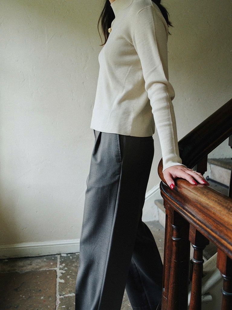 Woman wearing an eggshell high neck top in a fine rib with wide charcoal trousers on an oak staircase