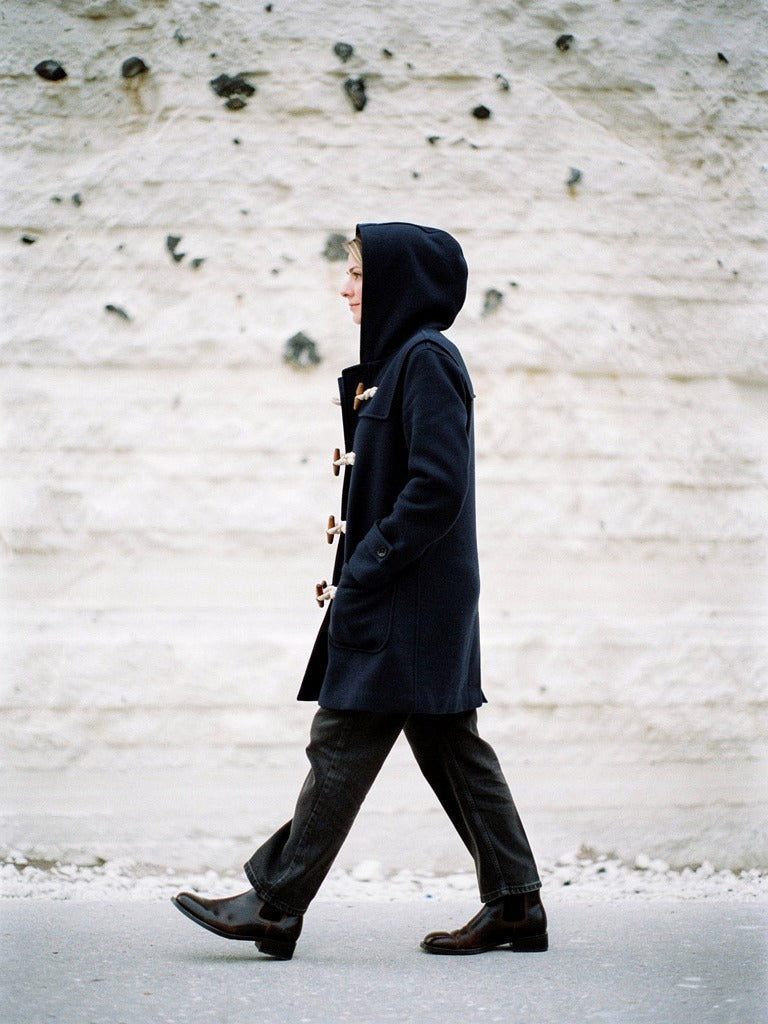 Navy hooded toggle coat worn with the hood up against a white chalk wall