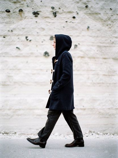 Navy hooded toggle coat worn with the hood up against a white chalk wall